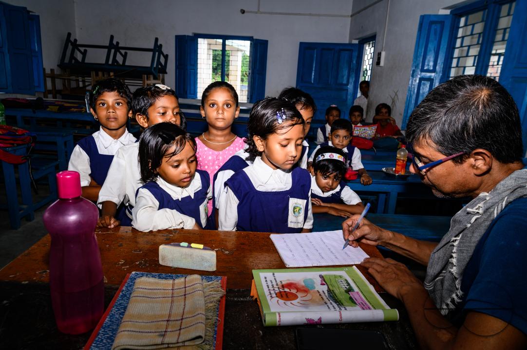At a rural school on the India- Bangladesh border side, a teacher is trying to explain to his class of pre-primary students (ages between 5 to 6 years) how to write English letters, students are constantly making fun of their teacher, and some are drawin