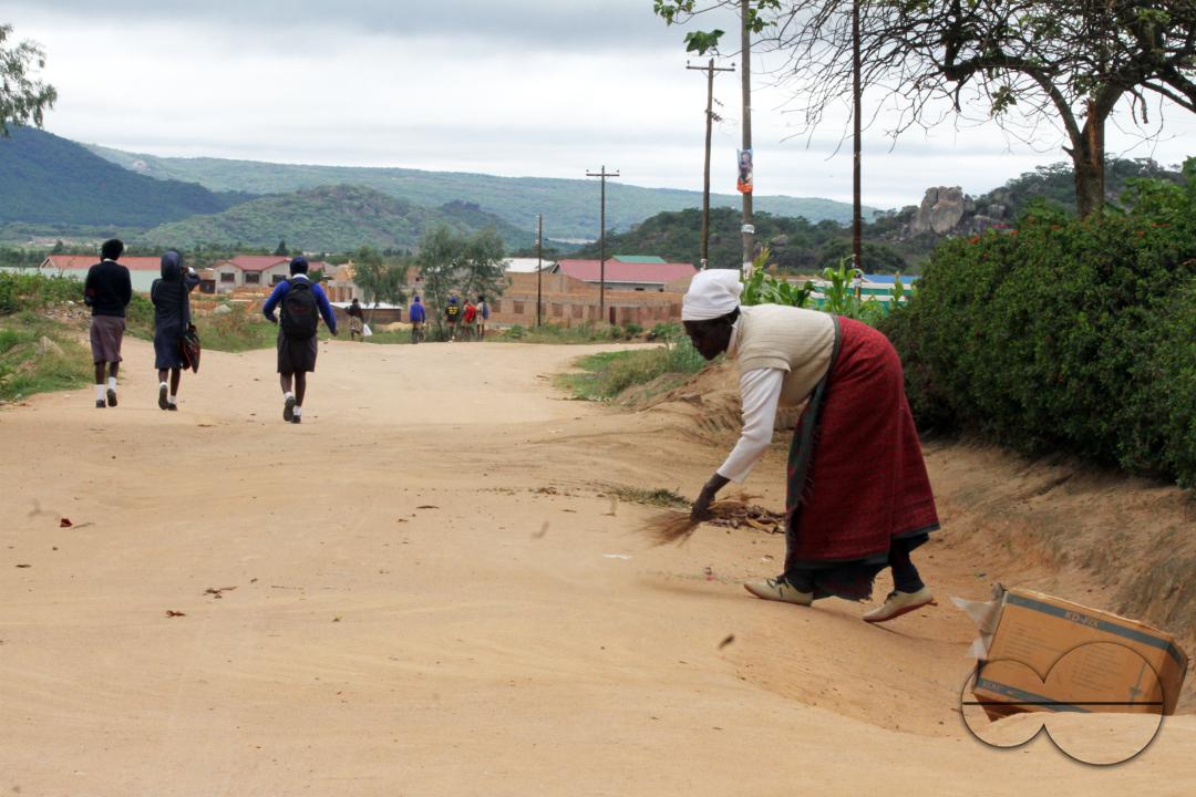 A woman is seen sweeping outside her yard early in the morning as scholars in uniform pass her  in Macheke township, Macheke