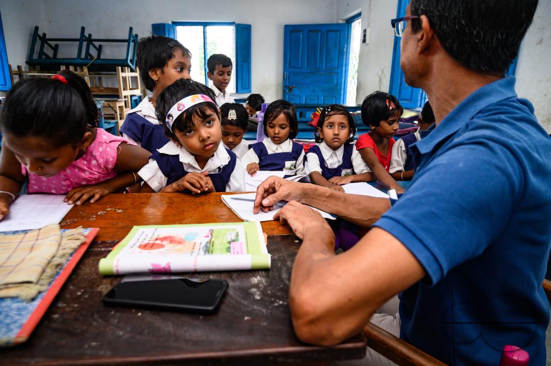At a rural school on the India- Bangladesh border side, a teacher is trying to explain to his class of pre-primary students (ages between 5 to 6 years) how to write English letters, students are constantly making fun of their teacher, and some are drawin