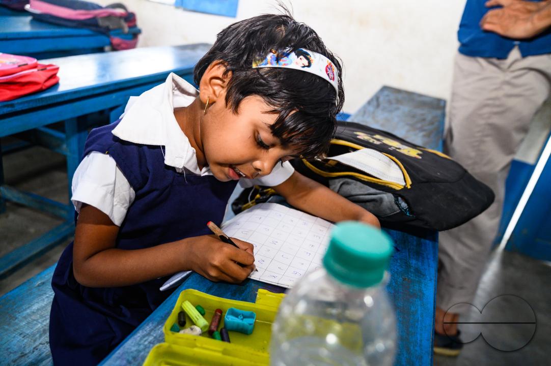 At a rural school on the India- Bangladesh border side, a teacher is trying to explain to his class of pre-primary students (ages between 5 to 6 years) how to write English letters, students are constantly making fun of their teacher, and some are drawin