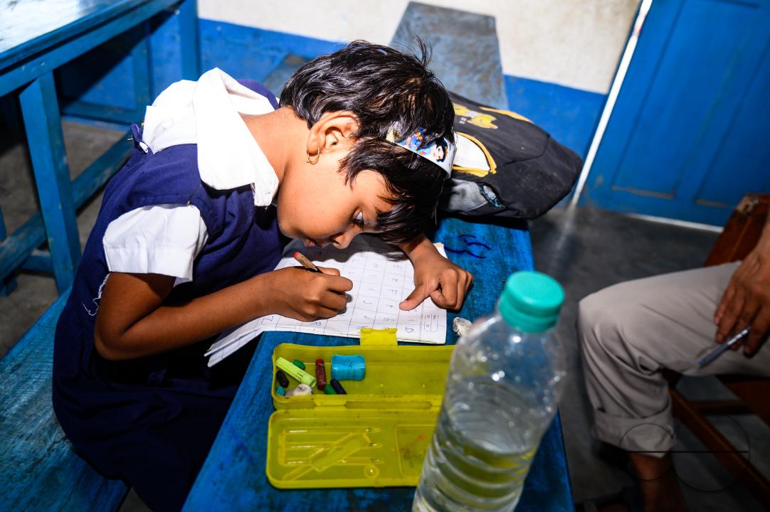 At a rural school on the India- Bangladesh border side, a teacher is trying to explain to his class of pre-primary students (ages between 5 to 6 years) how to write English letters, students are constantly making fun of their teacher, and some are drawin