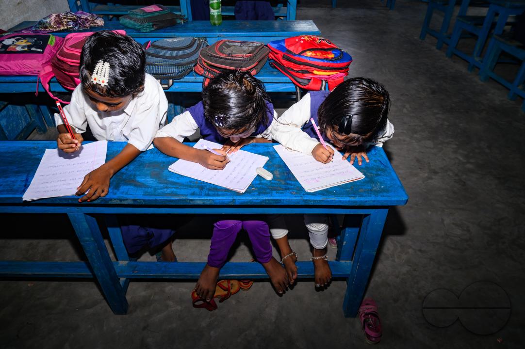 At a rural school on the India- Bangladesh border side, a teacher is trying to explain to his class of pre-primary students (ages between 5 to 6 years) how to write English letters, students are constantly making fun of their teacher, and some are drawin