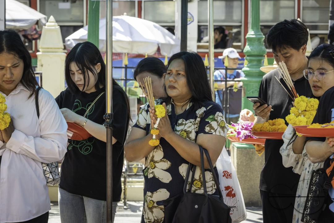 In downtown Bangkok, a diverse gathering of both Thai locals and tourists converge at the Erawan Shrine, each engaged in prayer with their respective offerings