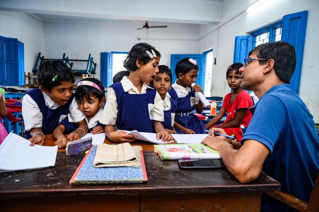 At a rural school on the India- Bangladesh border side, a teacher is trying to explain to his class of pre-primary students (ages between 5 to 6 years) how to write English letters, students are constantly making fun of their teacher, and some are drawin