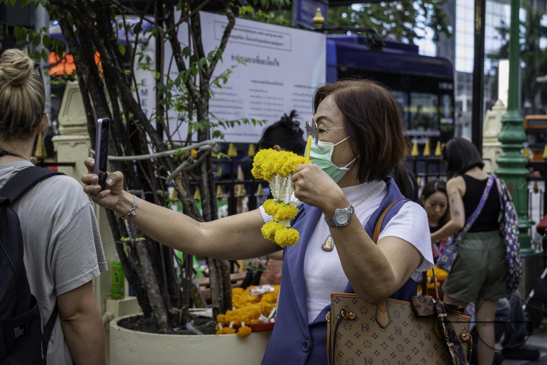At the Erawan Shrine in downtown Bangkok, an elderly Asian woman is taking a selfie with her flower garland, purchased for an offering